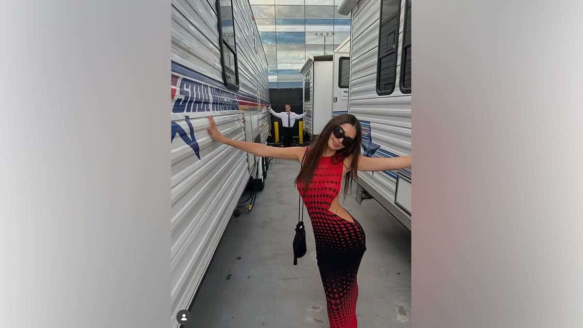 Emily Ratajkowski poses between production trailers while wearing her red-and-black VMAs look and sunglasses.