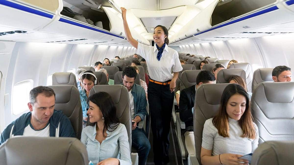 Flight attendant walks down airplane aisle closing overhead compartments and smiling.