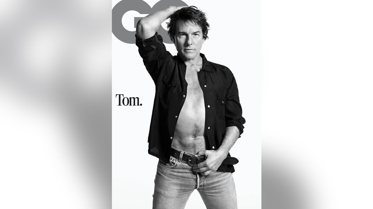 Tom Cruise poses with one arm raised while wearing an open dark shirt and jeans.
