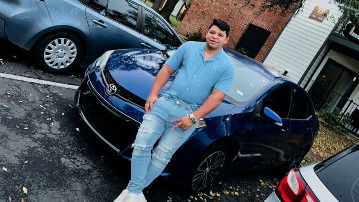 Wilber Garces in blue shirt and jeans posing in front of car