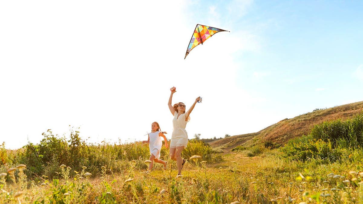 Woman and teenage girl run across a field while flying a kite.