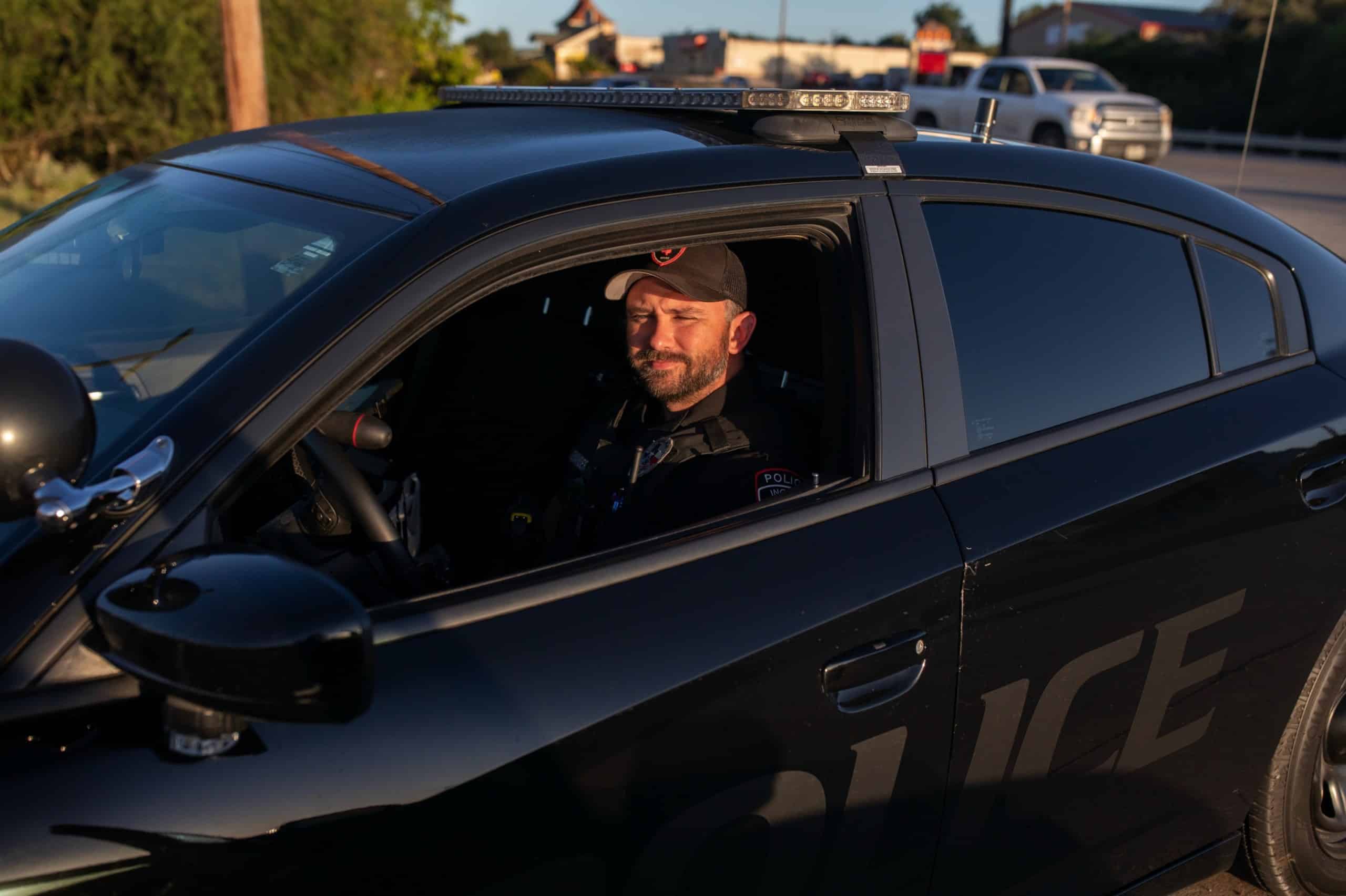 A bearded man wearing a baseball cap and a dark uniform sits in a black Charger.