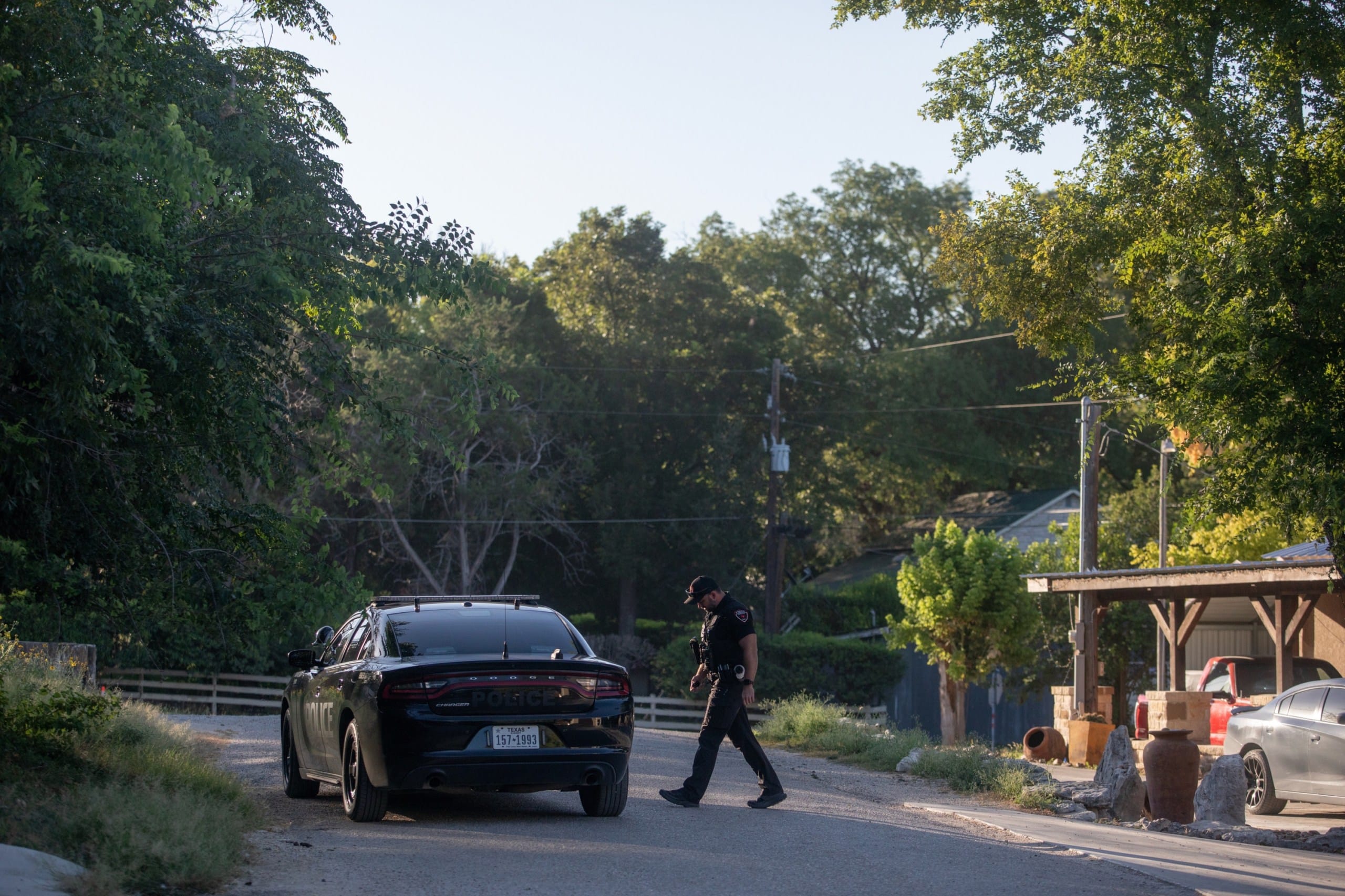 An officer, wearing a black uniform and a ballcap, crosses the street in a residential setting toward his parked Charger.