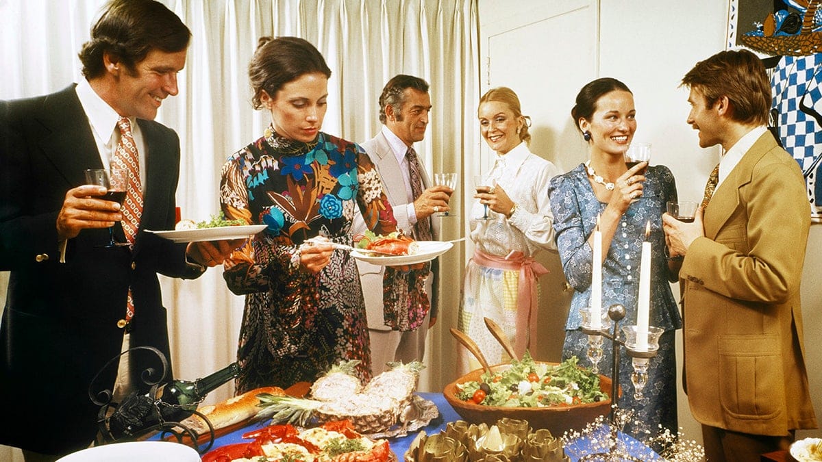 1970s Men And Women Around A Party Buffet Table Talking Drinking Wine Table Set With Salad Artichokes Lobster Roast Beef Fondue. (Photo by Photo Media/ClassicStock/Getty Images)