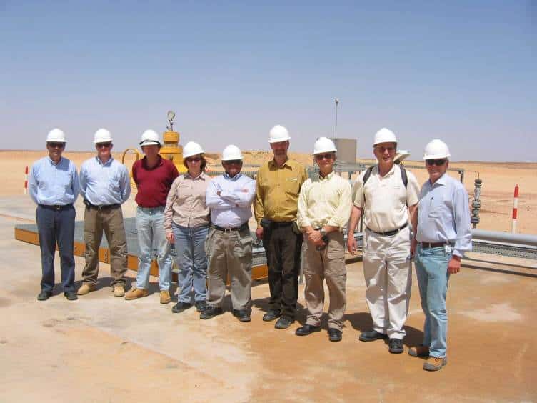Nine people wearing hard hats, sunglasses and field clothes stand in a line at an industrial energy site in a desert landscape.