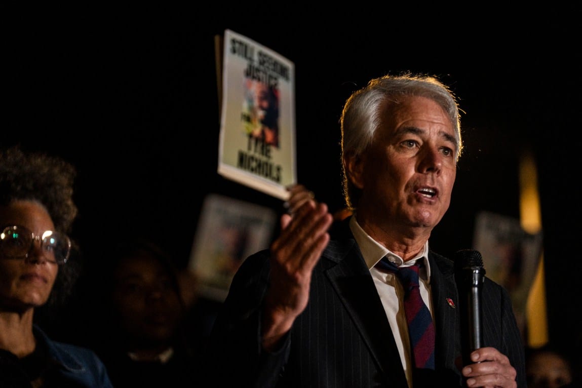 A man holding a microphone gestures with his hand as he speaks. In the background, a crowd is holding signs that say, “Still Seeking Justice. Tyre Nichols.”