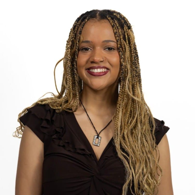 A woman with long blonde braids smiles at the camera and wears a black top and a necklace with a silver pendant.