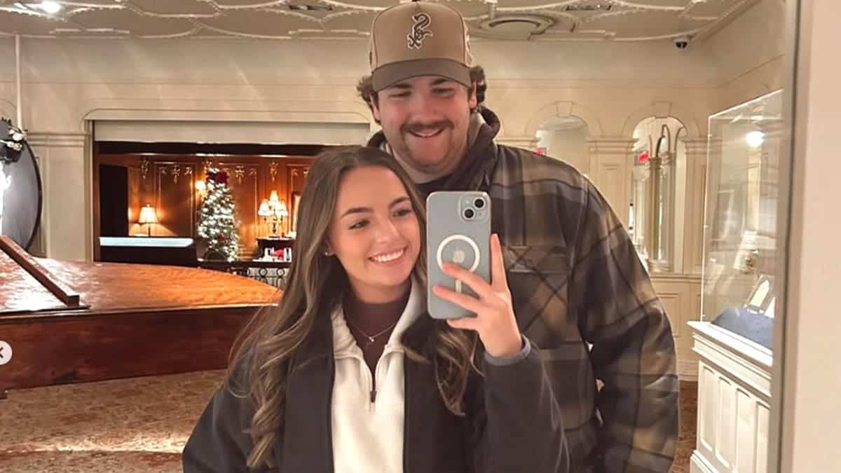 Leah Fonte smiles while taking a mirror selfie with her fiancé, firefighter Shane Hirstius, who stands behind her wearing a baseball cap and plaid jacket.
