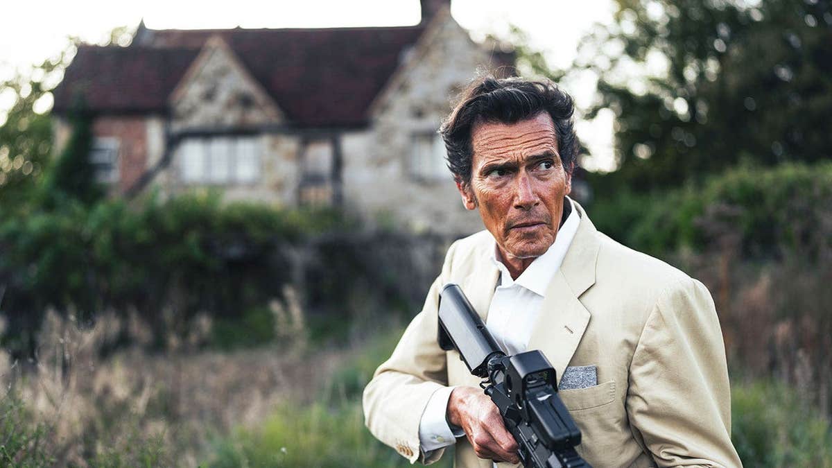 Geoffrey Moore in a white suit holding a gun outside an English cottage.