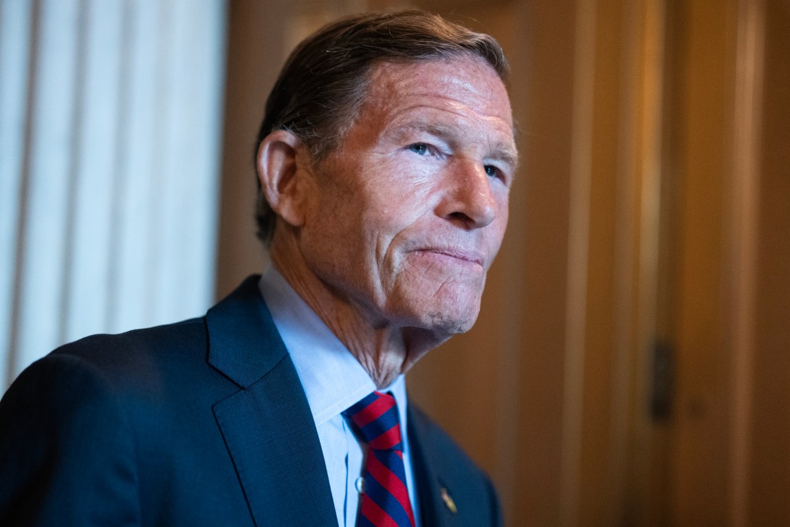 A man with brown hair wearing a blue suit and striped tie stands inside a room and looks off in the distance.