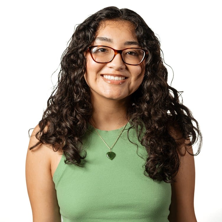 A woman with shoulder-length wavy dark brown hair smiles at the camera. She wears glasses, a green tank top and a silver necklace.