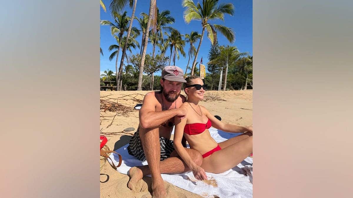 Kate Hudson wears a red strapless bikini and sunglasses while sitting beside her shirtless fiancé, Danny Fujikawa, on a sandy beach in Hawaii.