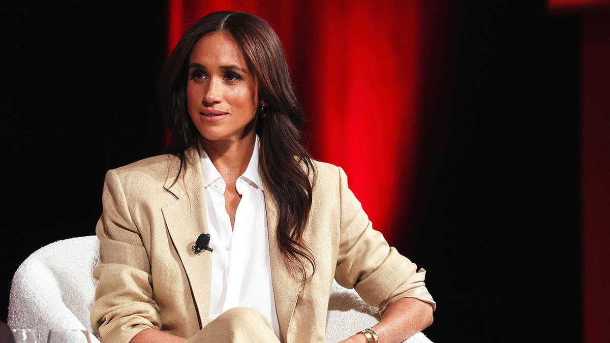Meghan Markle speaking onstage in a tan suit and white shirt.