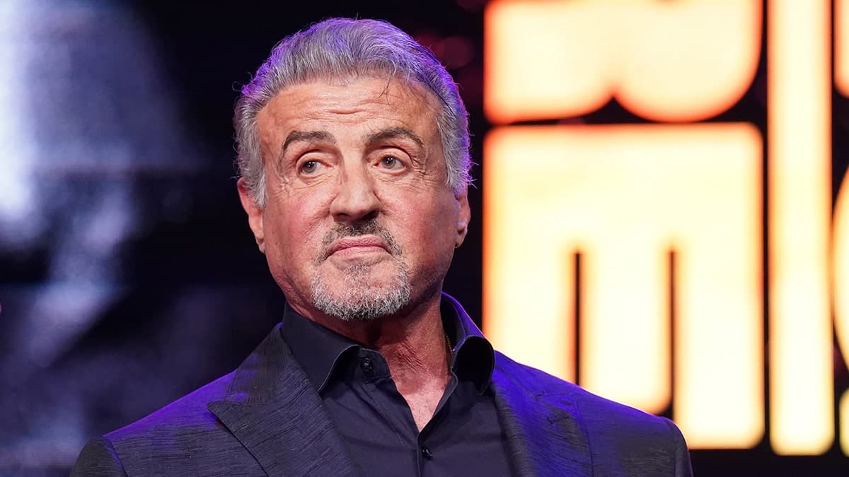 Sylvester Stallone standing in front of a lighted background while wearing a dark suit.
