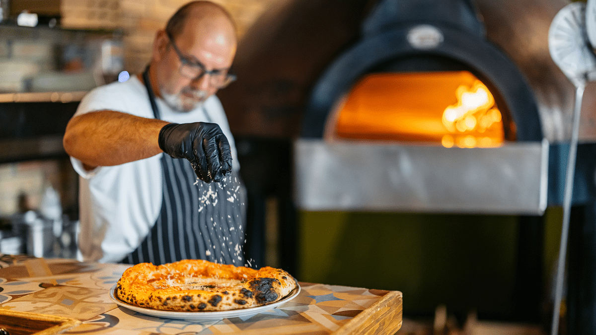 A mature chef garnishes a pizza after taking it out of the pizza oven in a commercial kitchen.