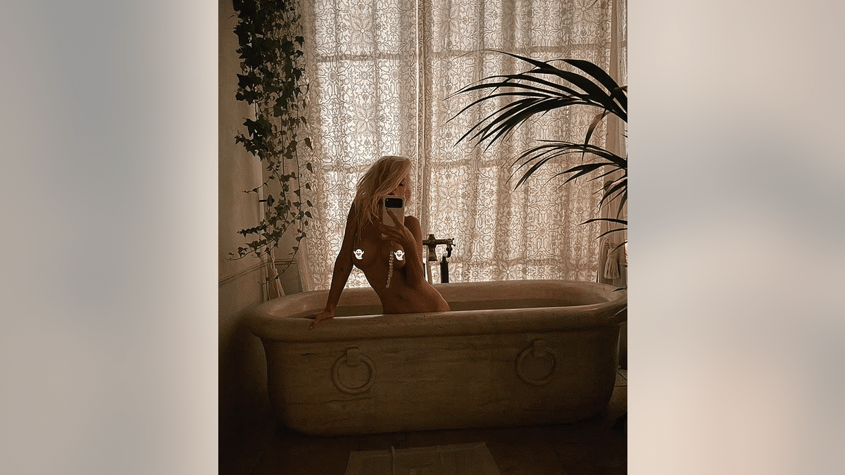 Christina Aguilera sits in a bathtub holding a smartphone in front of her face, with patterned curtains and plants behind her.