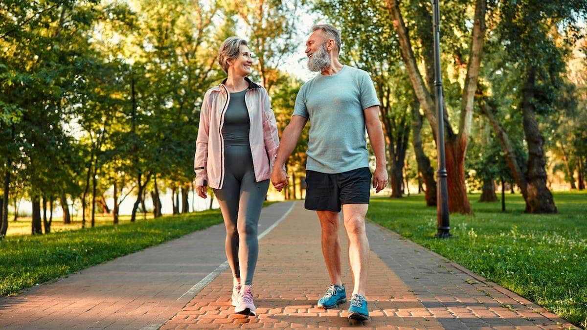 An elderly man and woman walk hand in hand in a park, looking at each other.