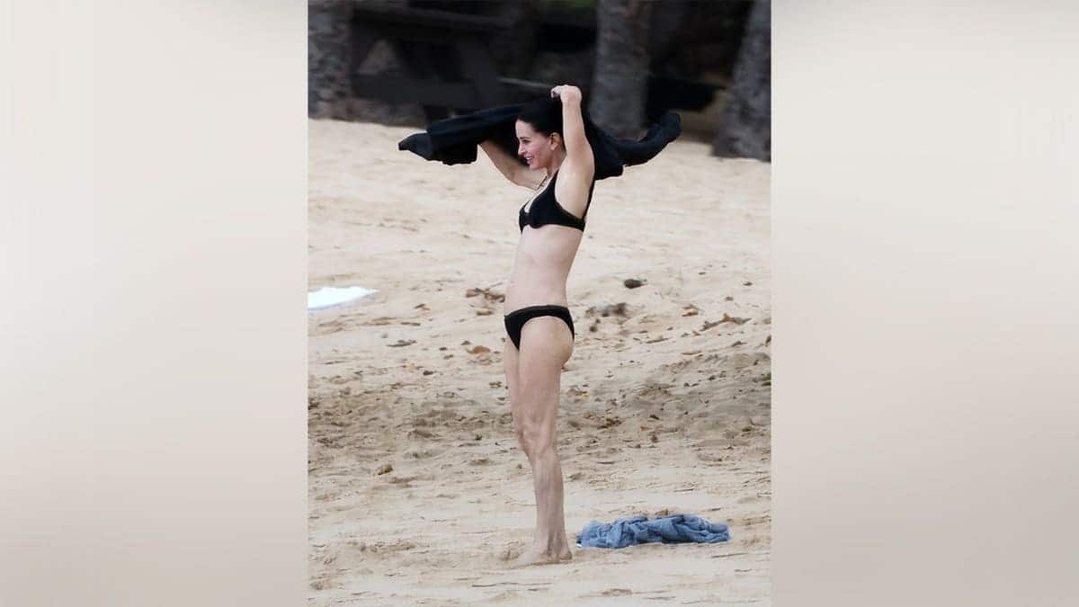 Courteney Cox standing on the beach in a black bikini.