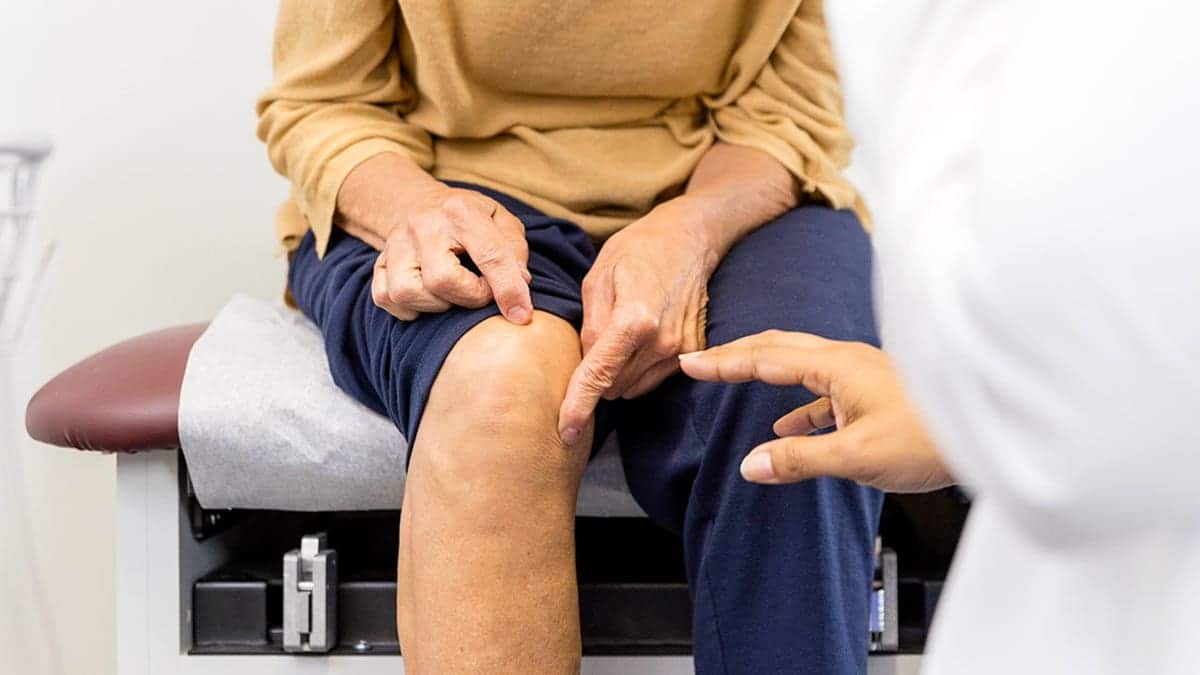 Senior woman sitting on examination table points to her knee while female doctor observes.