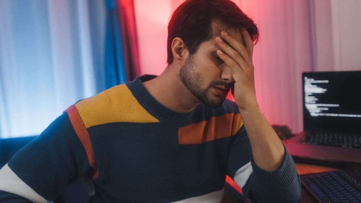 Tired man sits at desk with multiple computer screens in home office at night.