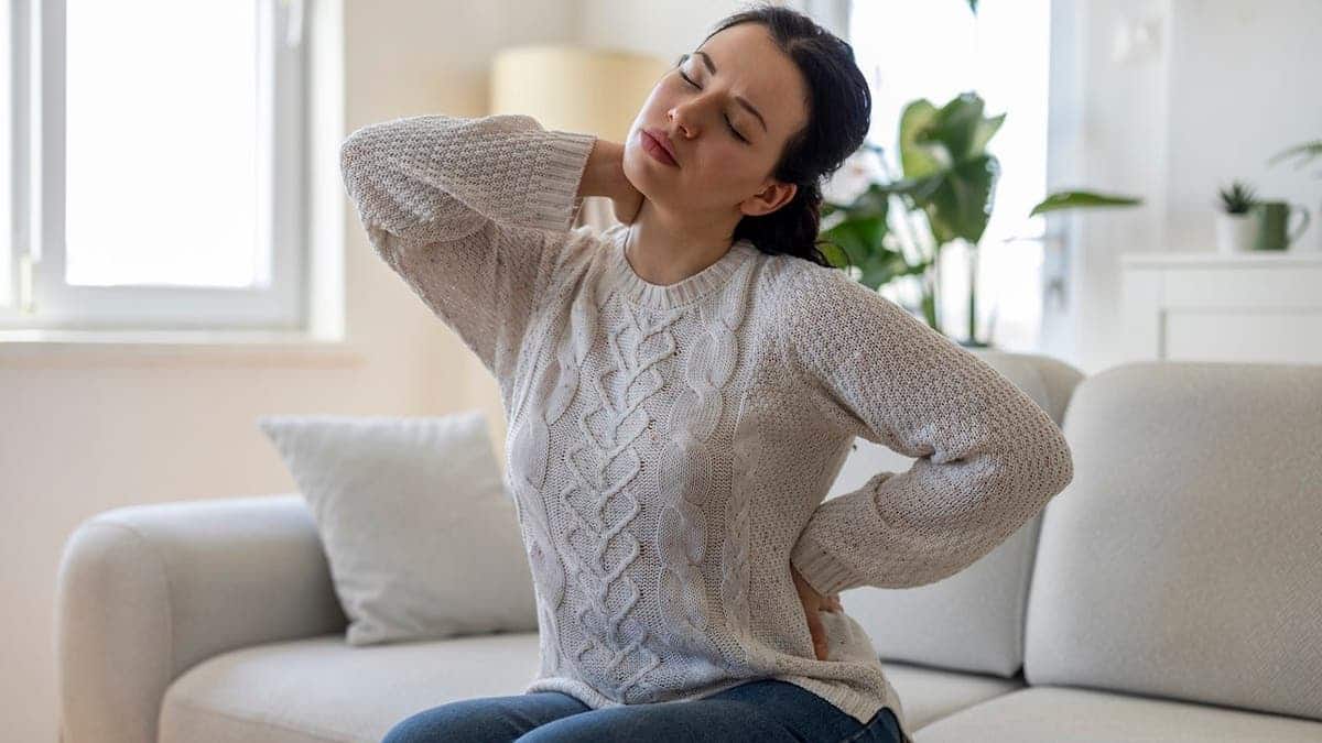Woman sitting on sofa, holding her neck and lower back in pain.