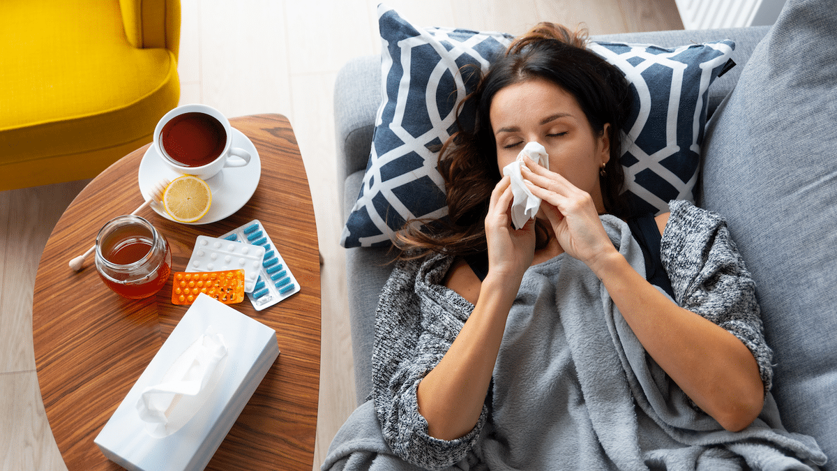 A woman lies on a couch with a tissue in her mouth while a cup of tea sits on a table next to her.
